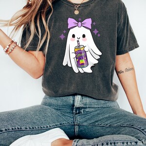 K&ouml;nnte beinhalten: Dunkelgraues T-Shirt mit einem Cartoon-Geist mit lila Schleife, der ein Getr&auml;nk h&auml;lt. Das Getr&auml;nk befindet sich in einer lila-gelben Dose mit dem Text "Alan's". Der Geist ist von lila Sternen umgeben.
