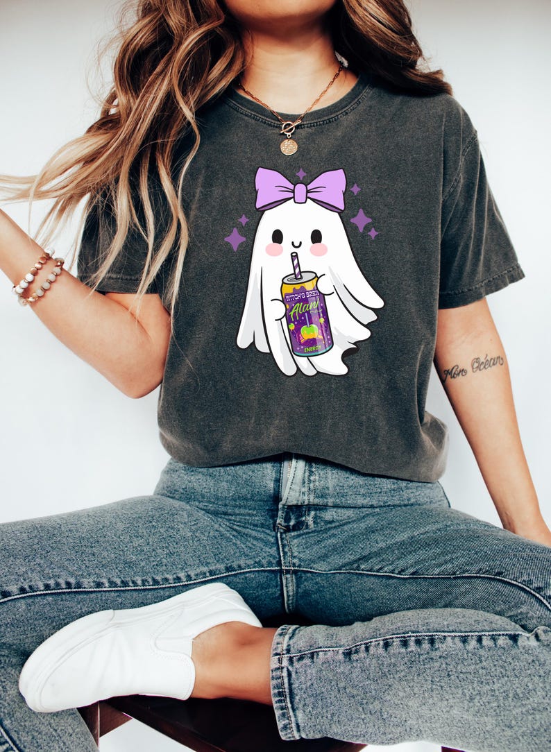 K&ouml;nnte beinhalten: Dunkelgraues T-Shirt mit einem Cartoon-Geist mit lila Schleife, der ein Getr&auml;nk h&auml;lt. Das Getr&auml;nk befindet sich in einer lila-gelben Dose mit dem Text "Alan's". Der Geist ist von lila Sternen umgeben.