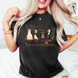 May include: Black t-shirt featuring a Halloween-themed graphic. The design includes four geese dressed in costumes: a ghost, a vampire, a pumpkin, and a skeleton. The text "just a spooky goose" is printed below the geese.