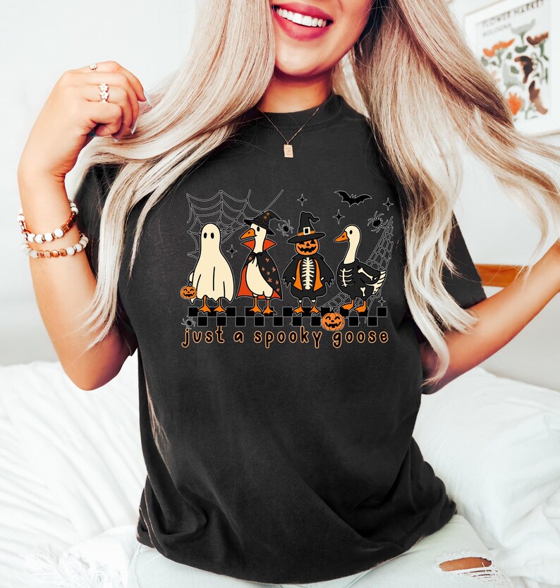 May include: Black t-shirt featuring a Halloween-themed graphic. The design includes four geese dressed in costumes: a ghost, a vampire, a pumpkin, and a skeleton. The text "just a spooky goose" is printed below the geese.