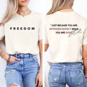 May include: Cream-colored t-shirt with the word "FREEDOM" printed in black on the front. The back of the shirt features the quote, "JUST BECAUSE YOU ARE OFFENDED DOESN'T MEAN YOU ARE RIGHT" in black and red text, with a signature.