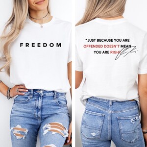 May include: White t-shirt with the word "FREEDOM" printed in black on the front. The back of the shirt features the quote, "JUST BECAUSE YOU ARE OFFENDED DOESN'T MEAN YOU ARE RIGHT" in black and red text, with a signature.