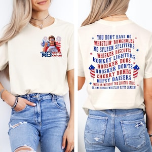 Pode incluir: Camiseta creme com a palavra "'MERICA" em vermelho, branco e azul, com um gr&aacute;fico de uma mulher usando &oacute;culos de sol e fogos de artif&iacute;cio. A parte de tr&aacute;s da camisa tem uma lista de frases humor&iacute;sticas.