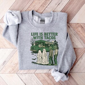 May include: A heather gray sweatshirt with a crew neck. The front features a green taco truck graphic with the text "LIFE IS BETTER WITH TACOS". Ghosts and zombies are also in the graphic.
