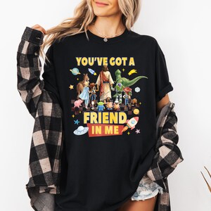 May include: A black short-sleeved t-shirt featuring a graphic print of various cartoon characters and a central figure walking together. The text "YOU'VE GOT A FRIEND IN ME" is printed in yellow and red. The shirt is layered under a black and tan plaid shirt.