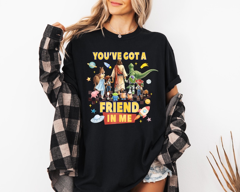 May include: A black short-sleeved t-shirt featuring a graphic print of various cartoon characters and a central figure walking together. The text "YOU'VE GOT A FRIEND IN ME" is printed in yellow and red. The shirt is layered under a black and tan plaid shirt.