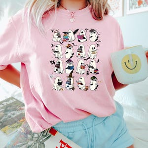 May include: Pale pink t-shirt with a pattern of cartoon ghosts reading books, wearing hats, and holding bats. The ghosts have black eyes and are surrounded by small bats and stars. The shirt is a light pink colour.