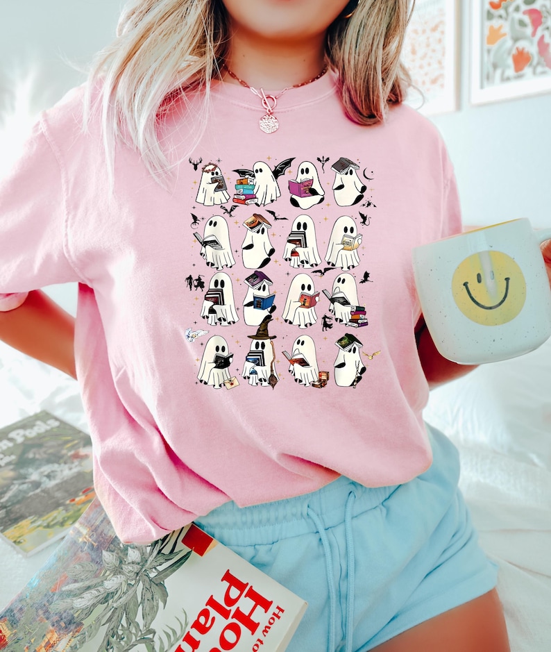 May include: Pale pink t-shirt with a pattern of cartoon ghosts reading books, wearing hats, and holding bats. The ghosts have black eyes and are surrounded by small bats and stars. The shirt is a light pink colour.