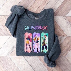 May include: A dark gray sweatshirt featuring the word "HUNTRIX" in a colorful, gradient font. Below the text are three anime-style illustrations of women in various outfits. The sweatshirt is folded on a light brown and white wooden surface.