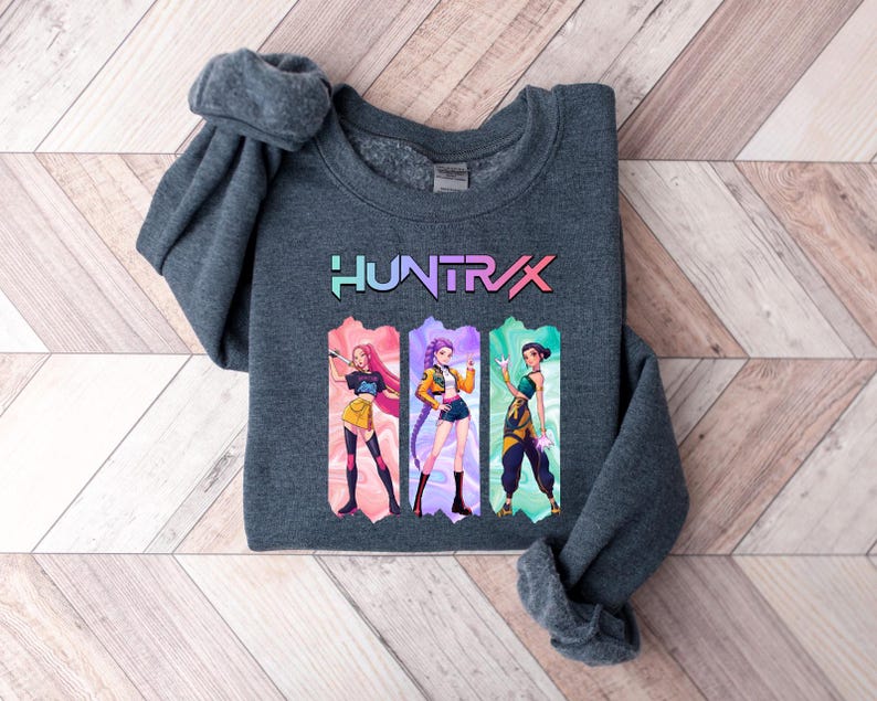 May include: A dark gray sweatshirt featuring the word "HUNTRIX" in a colorful, gradient font. Below the text are three anime-style illustrations of women in various outfits. The sweatshirt is folded on a light brown and white wooden surface.