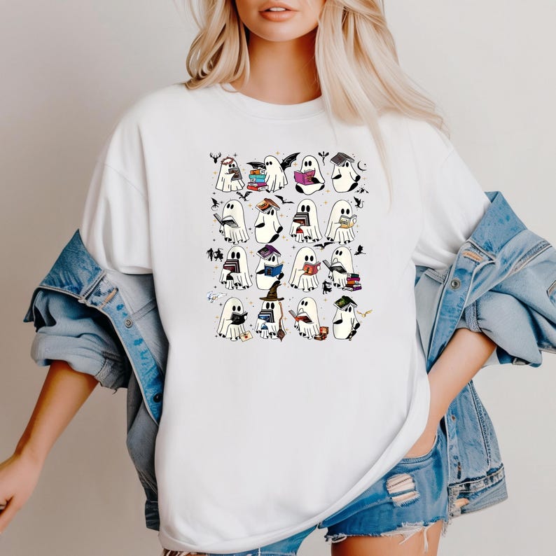 May include: White t-shirt with a graphic of cartoon ghosts reading books, wearing mortarboards, and holding witch hats. The design includes bats and stars, creating a Halloween or academic theme.