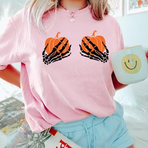 May include: A light pink t-shirt with a Halloween design. The design features two orange pumpkins held by black skeleton hands. The t-shirt has a crew neck.