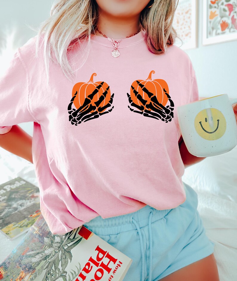 May include: A light pink t-shirt with a Halloween design. The design features two orange pumpkins held by black skeleton hands. The t-shirt has a crew neck.
