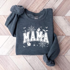 Op de afbeelding: Donkergrijze sweatshirt met het woord "MAMA" in witte blokletters, versierd met Halloween-thema graphics. Het ontwerp bevat spinnenwebben, vleermuizen, geesten en spinnen. De sweatshirt ligt op een licht houtnerf oppervlak.