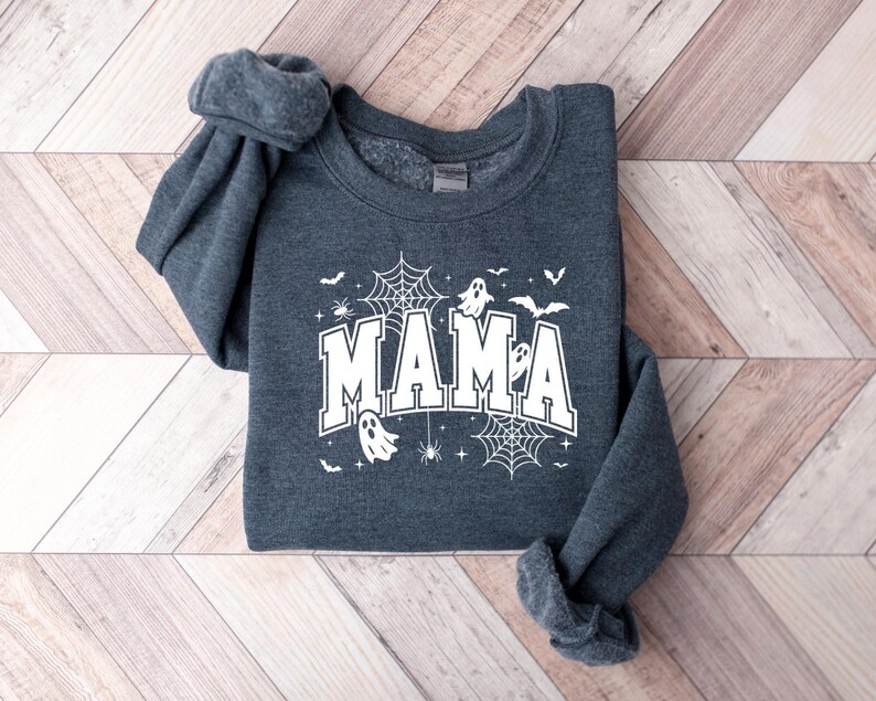 Op de afbeelding: Donkergrijze sweatshirt met het woord "MAMA" in witte blokletters, versierd met Halloween-thema graphics. Het ontwerp bevat spinnenwebben, vleermuizen, geesten en spinnen. De sweatshirt ligt op een licht houtnerf oppervlak.
