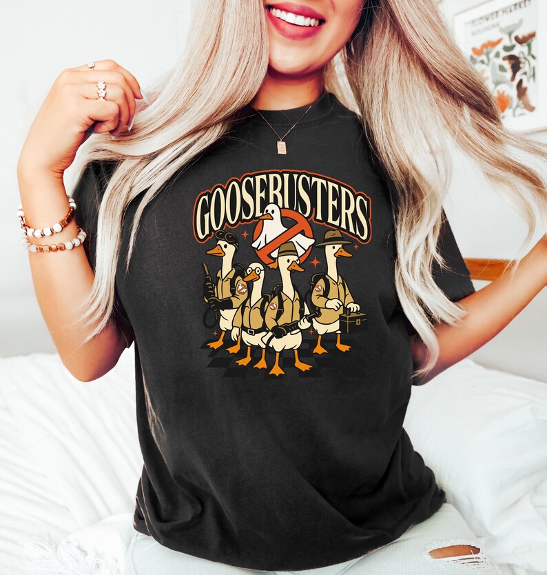 May include: Black t-shirt featuring a cartoon design of geese dressed as Ghostbusters, with the text "GOOSEBUSTERS" in an orange and white font. The geese are wearing khaki uniforms and carrying equipment. The design includes a ghost symbol.