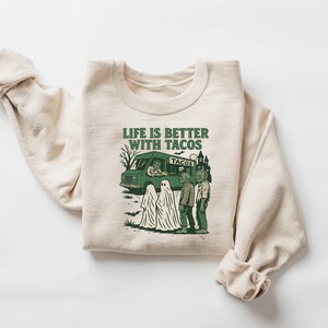 May include: Cream-colored sweatshirt with a green graphic that reads "LIFE IS BETTER WITH TACOS." The graphic features a taco truck, ghosts, and zombies. A fun, spooky design for Halloween or any day.