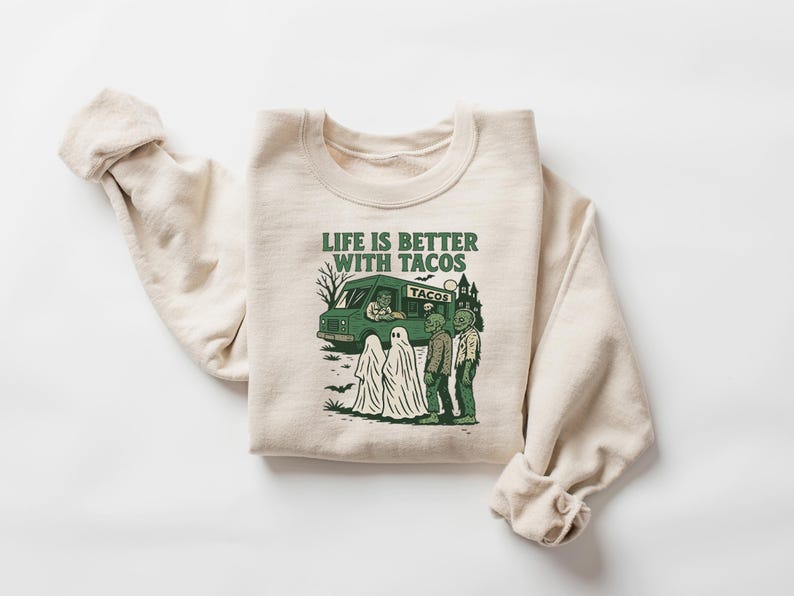 May include: Cream-colored sweatshirt with a green graphic that reads "LIFE IS BETTER WITH TACOS." The graphic features a taco truck, ghosts, and zombies. A fun, spooky design for Halloween or any day.