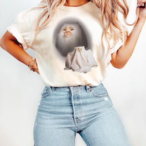 Puede incluir: Camiseta color crema con un dise&ntilde;o de gato. El dise&ntilde;o incluye la cara de un gato y un gato disfrazado de fantasma. El gato tiene pelaje naranja y blanco. La camiseta se combina con unos vaqueros azul claro.