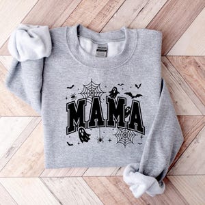 Op de afbeelding: Een lichtgrijs sweatshirt met het woord "MAMA" in zwarte blokletters, versierd met Halloween-thema graphics. Het ontwerp bevat spinnenwebben, vleermuizen, spinnen en geesten. Het sweatshirt ligt plat op een houten oppervlak.