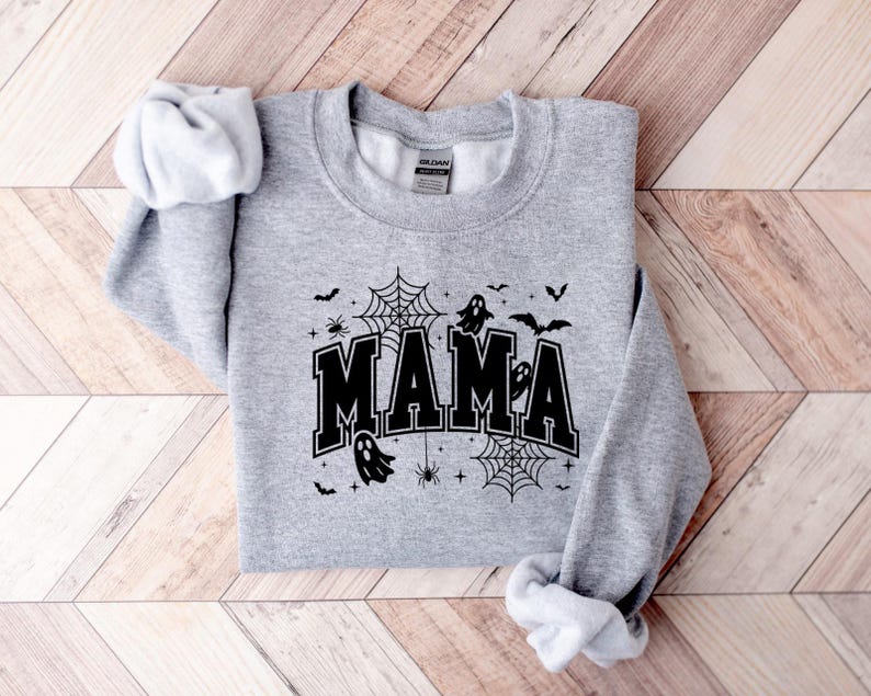 Op de afbeelding: Een lichtgrijs sweatshirt met het woord "MAMA" in zwarte blokletters, versierd met Halloween-thema graphics. Het ontwerp bevat spinnenwebben, vleermuizen, spinnen en geesten. Het sweatshirt ligt plat op een houten oppervlak.