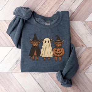 May include: A dark blue sweatshirt featuring a Halloween-themed graphic. The design includes three Highland cows dressed as a witch, a ghost, and a jack-o'-lantern. The witch cow wears a black hat and cape, while the pumpkin cow has a carved face.