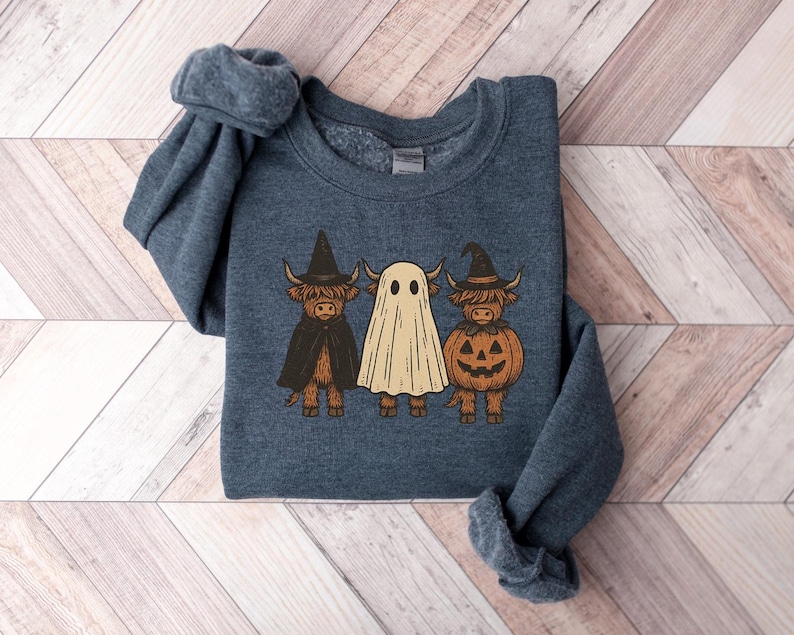 May include: A dark blue sweatshirt featuring a Halloween-themed graphic. The design includes three Highland cows dressed as a witch, a ghost, and a jack-o'-lantern. The witch cow wears a black hat and cape, while the pumpkin cow has a carved face.
