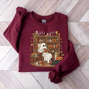 Peut inclure: Sweat-shirt bordeaux avec un motif sur le th&egrave;me d'Halloween. Le motif repr&eacute;sente un fant&ocirc;me lisant dans une biblioth&egrave;que confortable avec des citrouilles, des bougies et un chat noir. Le sweat-shirt est en mati&egrave;re douce.