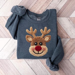May include: A dark blue sweatshirt featuring a reindeer design. The reindeer has brown antlers, a brown face, a red nose, and a tan mouth. The sweatshirt is made of a soft material and is perfect for the holiday season.