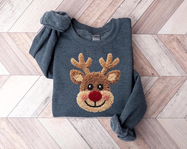 May include: A dark blue sweatshirt featuring a reindeer design. The reindeer has brown antlers, a brown face, a red nose, and a tan mouth. The sweatshirt is made of a soft material and is perfect for the holiday season.