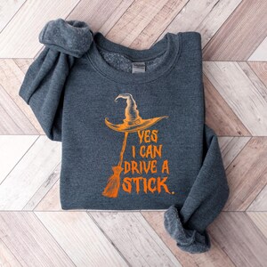 May include: Dark gray sweatshirt with the text "YES I CAN DRIVE A STICK" in orange. The graphic features a witch's hat and broom. The sweatshirt is folded on a light wood grain surface.