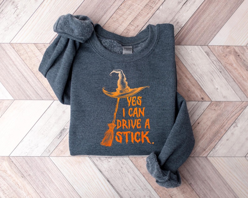 May include: Dark gray sweatshirt with the text "YES I CAN DRIVE A STICK" in orange. The graphic features a witch's hat and broom. The sweatshirt is folded on a light wood grain surface.