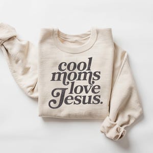 May include: A cream-colored sweatshirt with the phrase "cool moms love Jesus" printed in a dark, vintage-style font. The long sleeves are rolled up, and the sweatshirt is folded neatly on a white surface.