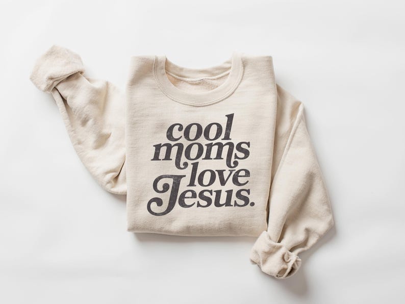 May include: A cream-colored sweatshirt with the phrase "cool moms love Jesus" printed in a dark, vintage-style font. The long sleeves are rolled up, and the sweatshirt is folded neatly on a white surface.