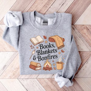 May include: A heather gray sweatshirt with the words "Books, Blankets & Bonfires" and illustrations of a book, blanket, bonfire, coffee mug, and marshmallow. The design features fall colors and cozy imagery.