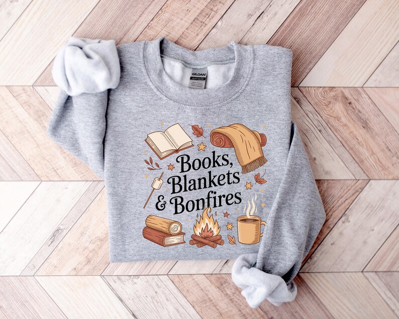 May include: A heather gray sweatshirt with the words "Books, Blankets & Bonfires" and illustrations of a book, blanket, bonfire, coffee mug, and marshmallow. The design features fall colors and cozy imagery.