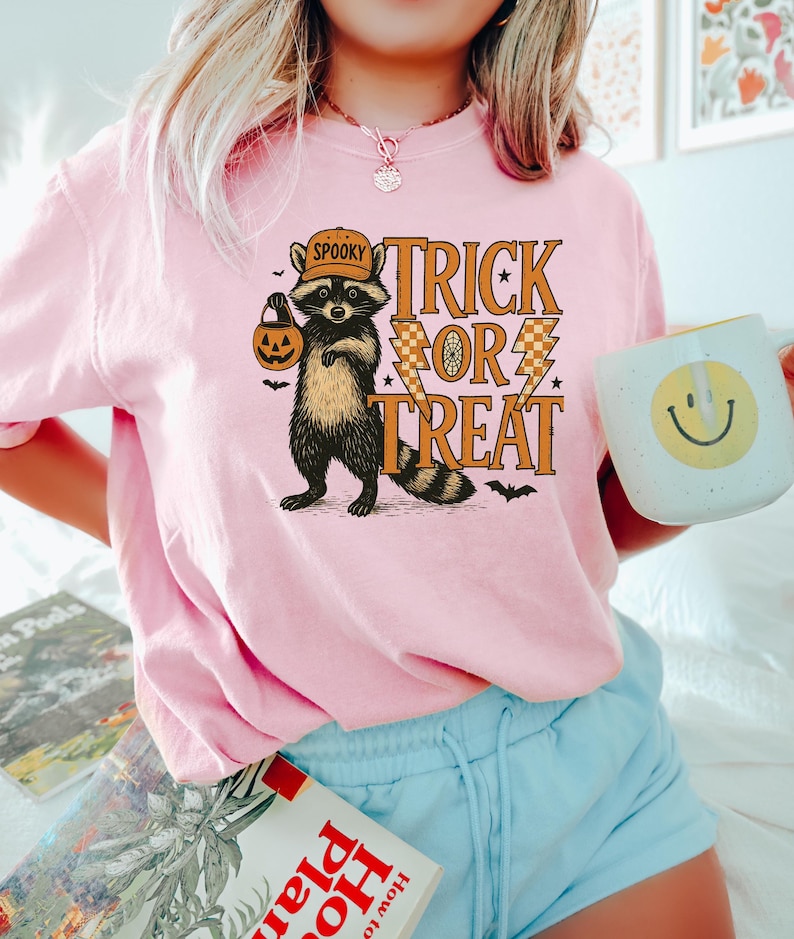 Peut inclure: Un t-shirt rose clair avec un motif d'Halloween. Le motif repr&eacute;sente un raton laveur portant un chapeau et tenant une lanterne, avec les mots "Trick or Treat". Une tasse blanche avec un smiley jaune est &eacute;galement visible.