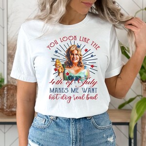 May include: White t-shirt with a red, white, and blue graphic of a woman holding a hot dog. The text on the shirt reads "You look like the 4th of July makes me want hot dog real bad."