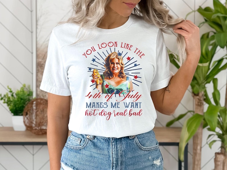 May include: White t-shirt with a red, white, and blue graphic of a woman holding a hot dog. The text on the shirt reads "You look like the 4th of July makes me want hot dog real bad."