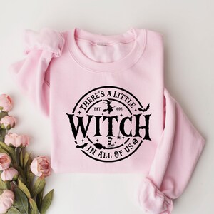 Op de afbeelding: Een lichtroze sweatshirt met de zwarte tekst "THERE'S A LITTLE WITCH IN ALL OF US" in een cirkel. Het ontwerp bevat een heks op een bezem, vleermuizen en sterren. Het sweatshirt ligt op een witte ondergrond met roze bloemen.