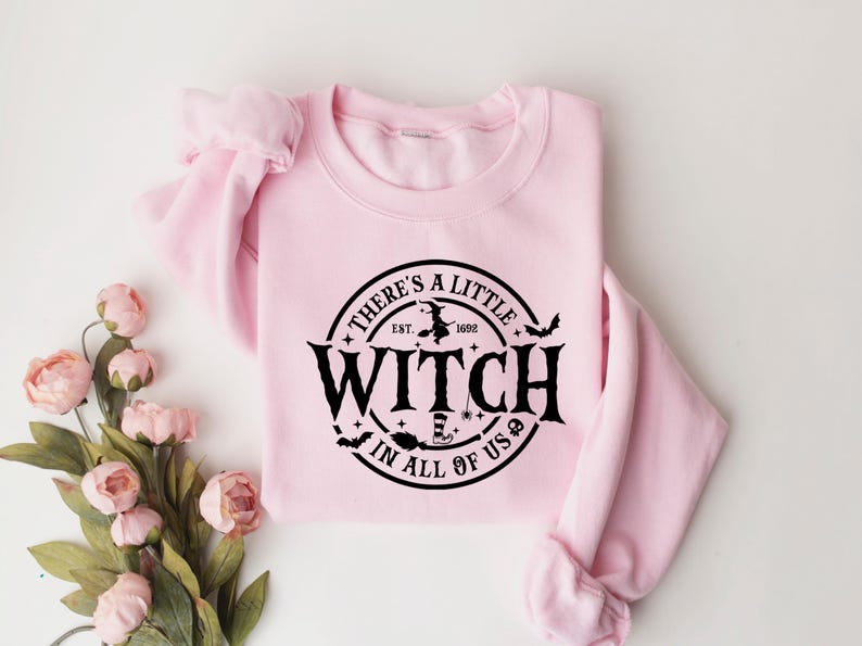 Op de afbeelding: Een lichtroze sweatshirt met de zwarte tekst "THERE'S A LITTLE WITCH IN ALL OF US" in een cirkel. Het ontwerp bevat een heks op een bezem, vleermuizen en sterren. Het sweatshirt ligt op een witte ondergrond met roze bloemen.