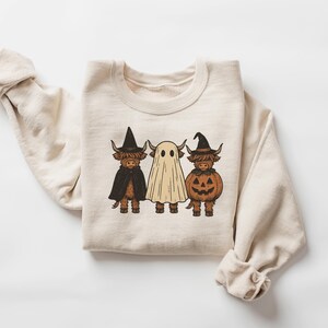 May include: Cream-colored sweatshirt featuring a Halloween-themed graphic. The design includes three Highland cows dressed as a witch, a ghost, and a jack-o'-lantern. The illustrations are in brown, black, and orange, perfect for the spooky season.