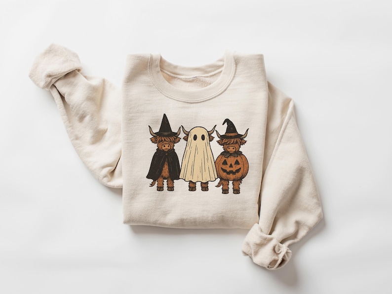 May include: Cream-colored sweatshirt featuring a Halloween-themed graphic. The design includes three Highland cows dressed as a witch, a ghost, and a jack-o'-lantern. The illustrations are in brown, black, and orange, perfect for the spooky season.