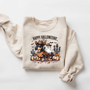 May include: Beige sweatshirt with a Halloween graphic. The design shows a dachshund in a cowboy hat and bandana, and a ghost wearing a saddle. The text "HAPPY HALLOWEENIE y'all" is above the image, with pumpkins and cacti.