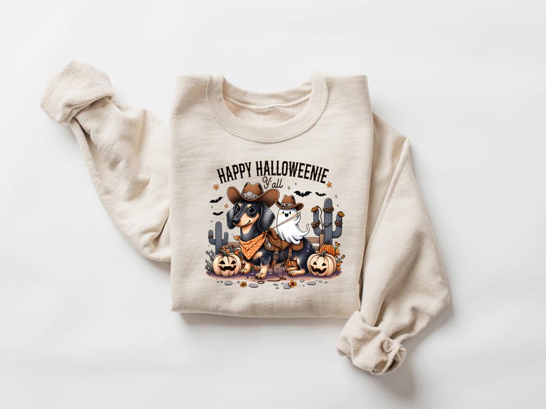 May include: Beige sweatshirt with a Halloween graphic. The design shows a dachshund in a cowboy hat and bandana, and a ghost wearing a saddle. The text "HAPPY HALLOWEENIE y'all" is above the image, with pumpkins and cacti.