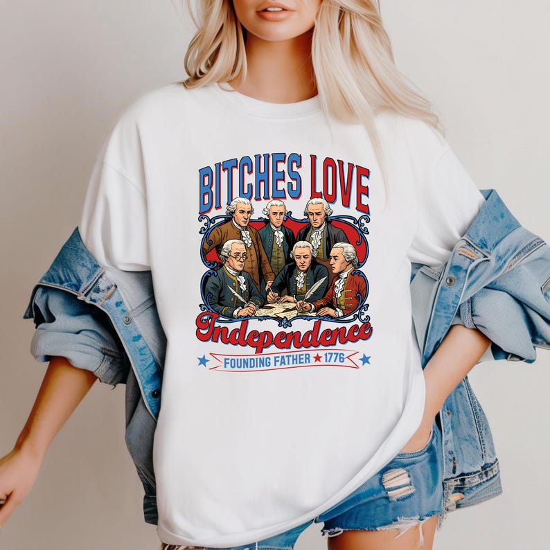 Może przedstawiać: Biała koszulka z grafiką z napisem "BITCHES LOVE Independence" w kolorze czerwonym i niebieskim. Grafika przedstawia grupę mężczyzn w strojach kolonialnych, z napisem "Founding Father 1776" poniżej.