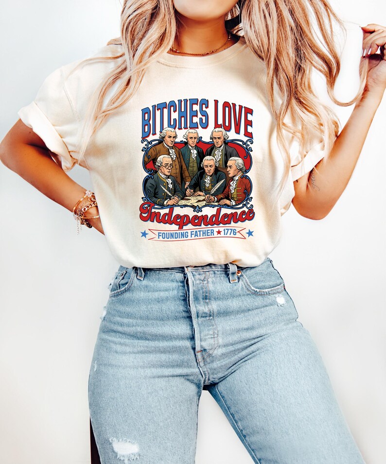 Może przedstawiać: Kremowa koszulka z nadrukiem graficznym. Tekst "BITCHES LOVE Independence" nad ilustracją Ojc&oacute;w Założycieli. Projekt zawiera czerwone, białe i niebieskie akcenty, z tekstem "FOUNDING FATHER 1776" poniżej.