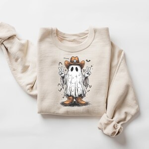 May include: Beige crewneck sweatshirt featuring a cartoon ghost wearing a cowboy hat and boots. The ghost is making peace signs with both hands. Bats and stars surround the ghost. A fun, whimsical design for Halloween.