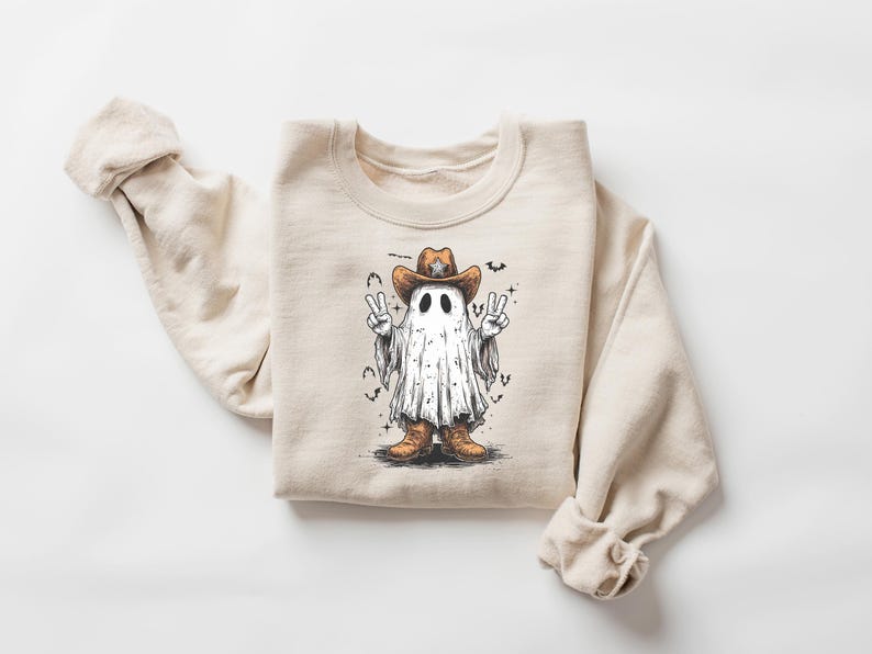 May include: Beige crewneck sweatshirt featuring a cartoon ghost wearing a cowboy hat and boots. The ghost is making peace signs with both hands. Bats and stars surround the ghost. A fun, whimsical design for Halloween.
