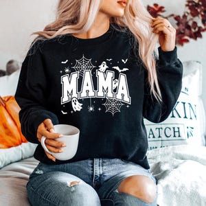 Op de afbeelding: Zwart sweatshirt met het woord "MAMA" in witte letters, versierd met spinnenwebben, vleermuizen, spinnen en geesten. Een ontwerp met Halloween-thema.